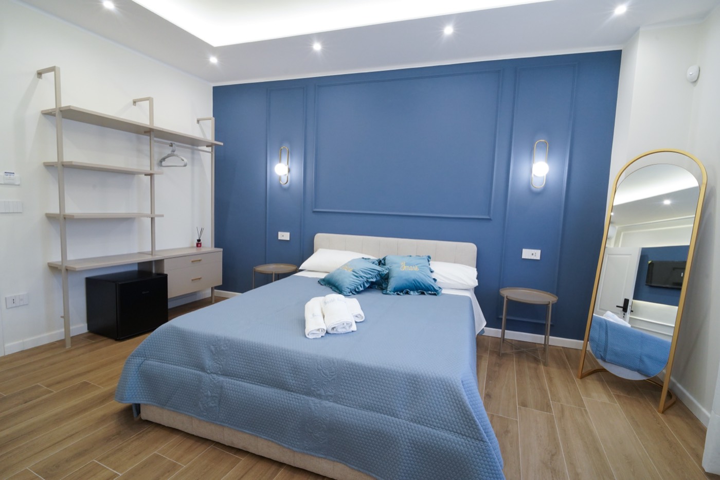Camera Blu: letto matrimoniale, parete blu con boiserie, libreria aperta con minibar e specchio ad arco dorato