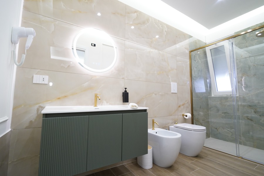 Bagno privato della Camera Orchidea con mobile verde, specchio tondo retroilluminato e rubinetteria dorata